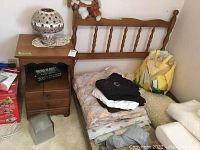 Wood and metal twin bed frame with stacked bedding, wood bedside table with ceramic lamp on top, and various items around