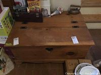 Top and front view of a rectangular wooden chest with a hinged lid and a heart-shaped cut-out on the front panel. Shows the chest among other items in a room.