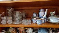 Shelf showing pinwheel cut crystal pieces including vases, candlesticks, bowls, and a glass vase along with a blue floral patterned ceramic teapot, cup, and saucer set plus cream and sugar bowls, and small white plates.