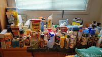Wide view of large assortment of cleaning supplies arranged on a table by a window, various spray bottles, powders, liquids.