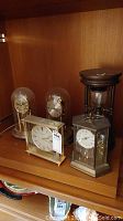 Four mantel clocks and one large wooden timer displayed on a wooden shelf. Clocks vary in shape including domed glass and rectangular forms.