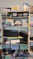 View of shelving unit containing multiple keyboards, a black radio, several plastic bins filled with cords and cables, white computer speakers, and stacks of blank CDs and small electronics boxes.