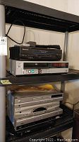 Image shows two shelves with various DVD players and VCRs including a black Panasonic VCR/DVD combo player on top and a silver JVC VCR below it.