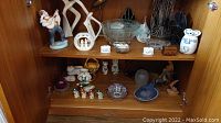 Two wooden shelves with assorted ceramic figures, candle holders, glass bowls, and trinket boxes arranged on them.