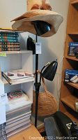 Black metal floor lamp with two adjustable lamp heads, with two straw hats placed on top. Woven straw handbag hanging from lamp stand. Shelves and books in background.