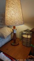 Photo of mid-century modern wood base table lamp with woven conical shade next to wood side table and glass miniature display cabinets.