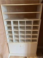 Full front view of the white storage shelf showing two open shelves on top, 24 small cubbies in the middle, and the pull-down shoe rack compartment at the bottom.