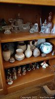 Wide view of three wooden shelves containing glassware, vases, pots, and small figurines arranged in rows