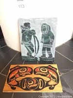 Front view of the marble plaque depicting two Inuit figures in traditional clothing and the Haida cedar wood plaque below