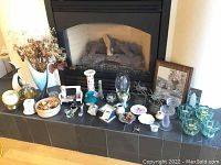 Wide view of the collection displayed on a black tiled hearth in front of a fireplace; includes framed art, glass candle holders, ceramics, and decorative items