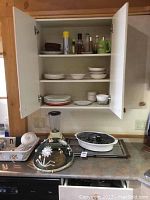 Kitchen cabinet with plates, bowls, bottles, cups, Hamilton Beach blender, wood cutting board, and Salton warming tray on counter