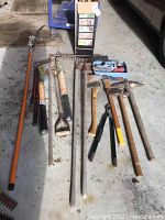 Wide view showing various garden tools laid out on the floor including pole trimmer, axes, maul axe, pickaxe, rakes, and other hand tools
