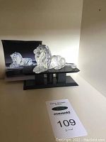 Clear crystal lion figurine displayed on black platform with black box in background showing image of the lion.