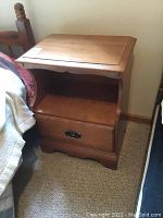 Wooden nightstand with a single drawer and open shelf placed beside a bed, viewed from side angle showing top surface and drawer.