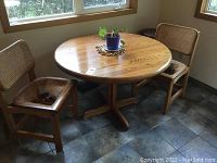 Solid wood round table with two wicker chairs placed on tiled floor near windows.