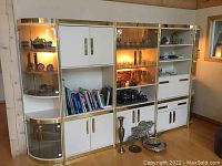 Front view of the entire large vintage wall unit showing glass shelves, gold framing, white storage cabinets, and lighting inside displays.