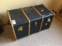 Front angled view of black trunk with wood straps, brass locks, and white chalk markings on top