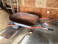 Full side view showing orange metal frame with black handle grips and single wheel, rust on metal tray.