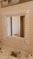 Square wall mirror with white iron frame made of undulating strips creating a decorative wave pattern.