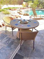 Outdoor round stone patio table with gas grill in center and two wicker chairs around it, set on tiled floor by pool.
