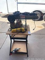 Wide photo showing Central Machinery belt and disc sander on stand with several corded power tools including a yellow hand drill and angle grinder on lower shelf.