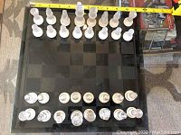 Top view showing full glass chessboard with all chess pieces arranged in starting positions. Chessboard features checkerboard pattern of frosted and clear glass squares. Pieces are clear and frosted glass.
