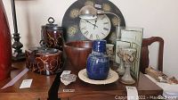 Overall view of wall clock, wooden bowl, blue vase, curved picture frames, brown containers, candle holder, and additional decor on wooden table.