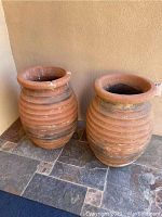 Photo showing two large red clay earthenware pots with ribbed design placed on a stone floor against a beige wall. One pot has visible damage on its rim.