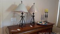 Two black wrought iron table lamps with beige fabric shades, two black wrought iron candle holders with yellow taper candles, and decorative wall platter above wooden dresser surface.