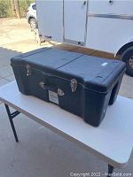 Large black plastic locker with metal clasps and handle on a white table outdoors, side view