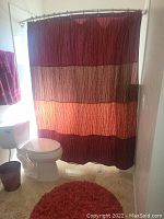 Photo showing bathroom with burgundy, orange, and tan striped shower curtain, matching burgundy rug, trash can, and towel, placed by white toilet.