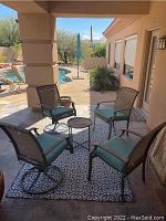 Four patio chairs with blue-green polyester batting cushions on a patterned blue-gray rug around a small round metal table with macramé top, outdoors on covered patio.