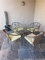 Angle showing square glass table top, ornate metal base, and four chairs with cushions including two metal design chairs and two wicker-style chairs