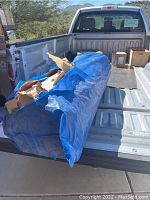 Blue tarp-covered large roll of upholstery fabric in truck bed with some torn paper wrapping indicating protection wrapping of sofa material.