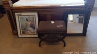 Overall view of padded stool, framed art print resting against bed base, and rectangular mirror.