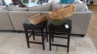 Photo showing two wooden stools, dark brown finished wood, with black leather cushioned seats. Basket items placed on stools include a rectangular wicker basket and a large rounded basket.
