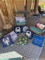Photo showing multiple ocean-themed throw pillows arranged on a patterned rug. Includes pillows with white lobster, white crab, white coral on blue backgrounds, and green palm leaf design.