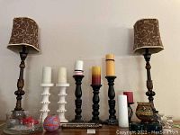 Two tall matching resin lamps with brown flocked cloth shades surrounded by an assortment of various candle holders and candles including wooden stands, glass candlesticks, a colored metal holder, multiple pillar and taper candles, and a glass vessel with pebbles and floating candles.