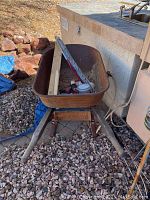 Metal wheelbarrow viewed outdoors on gravel near a patio area containing various items inside the wheelbarrow (contents not included). Wooden handles and a deflated tire visible.