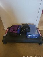 Side view of a black step-up platform with two purple yoga straps, a black mesh bag, and a blue yoga mat stacked on top.