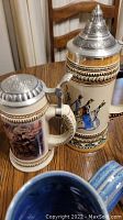 Beer steins on table showing a stein with metal lid and riders on horseback painted design