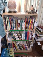 Front view of a light-colored laminate bookcase filled with books, showing three shelves with various books placed upright and some laying down on the bottom shelf. A lamp and figurine are on top of the bookcase.