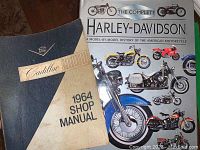 Photo showing the 1964 Cadillac Shop Manual with blue and beige cover and Cadillac branding alongside The Complete Harley-Davidson book with illustrated motorcycle cover.