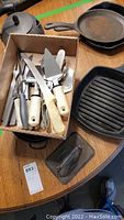Photo showing two cast iron pans, a cast iron grill pan, a box of assorted cutlery and kitchen utensils, and a metal nutcracker on a wooden table.