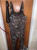 Front view of RedHead Mossy Oak Extreme Series camo fishing wader with attached suspenders hung on door.