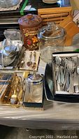 Table with vintage ceramic cookie jar labeled 'Cookies' with floral design and clear glass cookie jar with lid. Various flatware in silver and gold tones placed on trays. Wooden knife block with knives in background