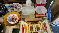 View of the combined lot table showing various colored Pyrex fridge boxes with lids, ceramic bowls with colorful artwork, green small dishes, wooden rolling pin, and ceramic tableware