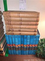 Photo showing a stack of vintage Hardy Boys and Franklin W. Dixon mystery books with tan and blue covers on wooden shelves.