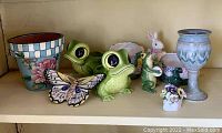 Shelf display of various garden-themed ceramic pottery items including frog planters, butterfly decoration, rabbit figurine, flower pots, and ceramic chalice.