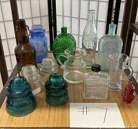 Photo shows the entire group of vintage glass bottles, insulators, and pitchers arranged on a wooden table for overview.